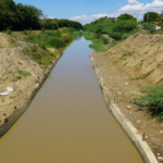 Canal con agua entre márgenes de tierra y vegetación, con maquinaria al fondo.