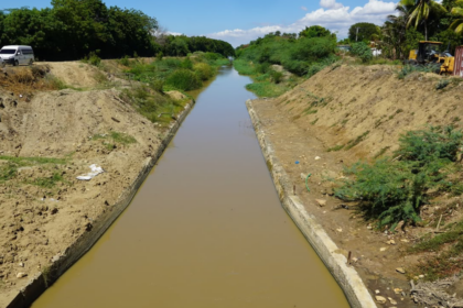 Canal con agua entre márgenes de tierra y vegetación, con maquinaria al fondo.