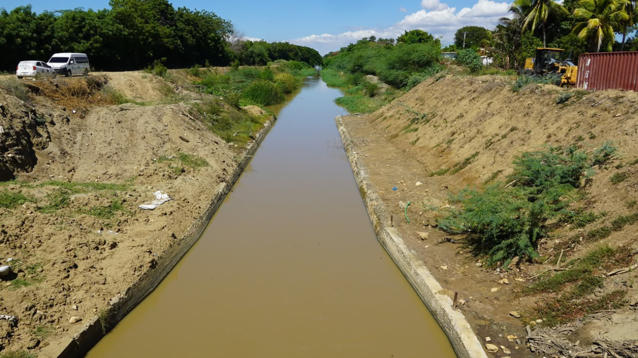 Canal con agua entre márgenes de tierra y vegetación, con maquinaria al fondo.