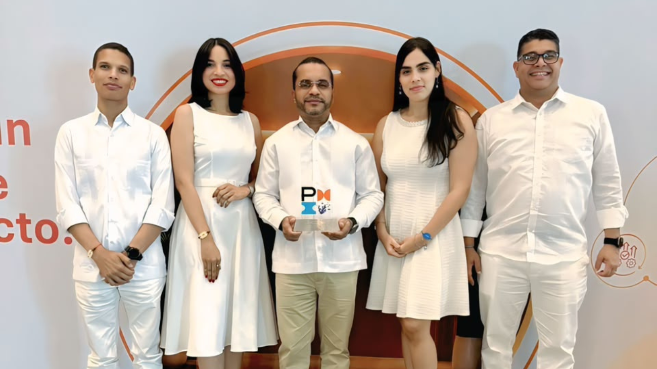 Cinco personas posan ante la gráfica de un evento; una sostiene un trofeo con la marca PMI.