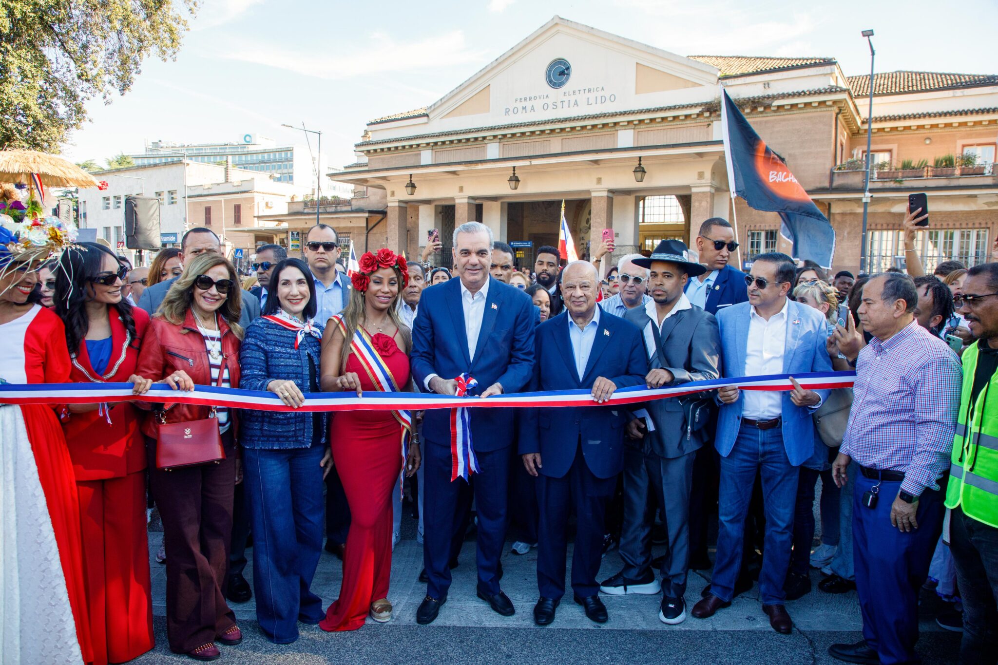 Participantes junto a una cinta roja, blanca y azul, con banderas y un edificio al fondo.