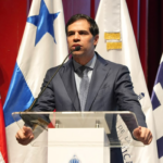 Ponente ante un atril y varias banderas durante una actividad institucional.