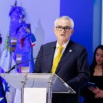 Orador en un atril con banderas y señalética institucional.