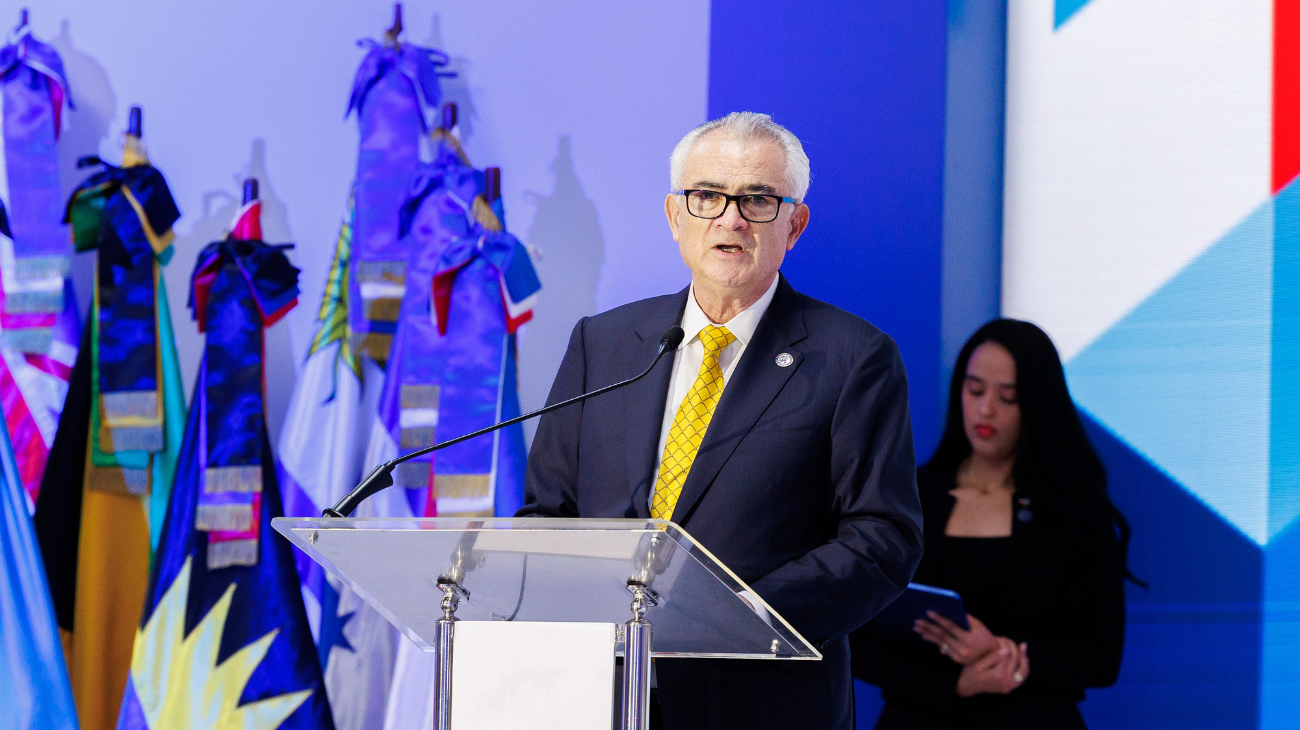 Orador en un atril con banderas y señalética institucional.