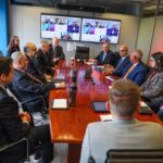 Participantes alrededor de una mesa con banderas y pantallas de videoconferencia.