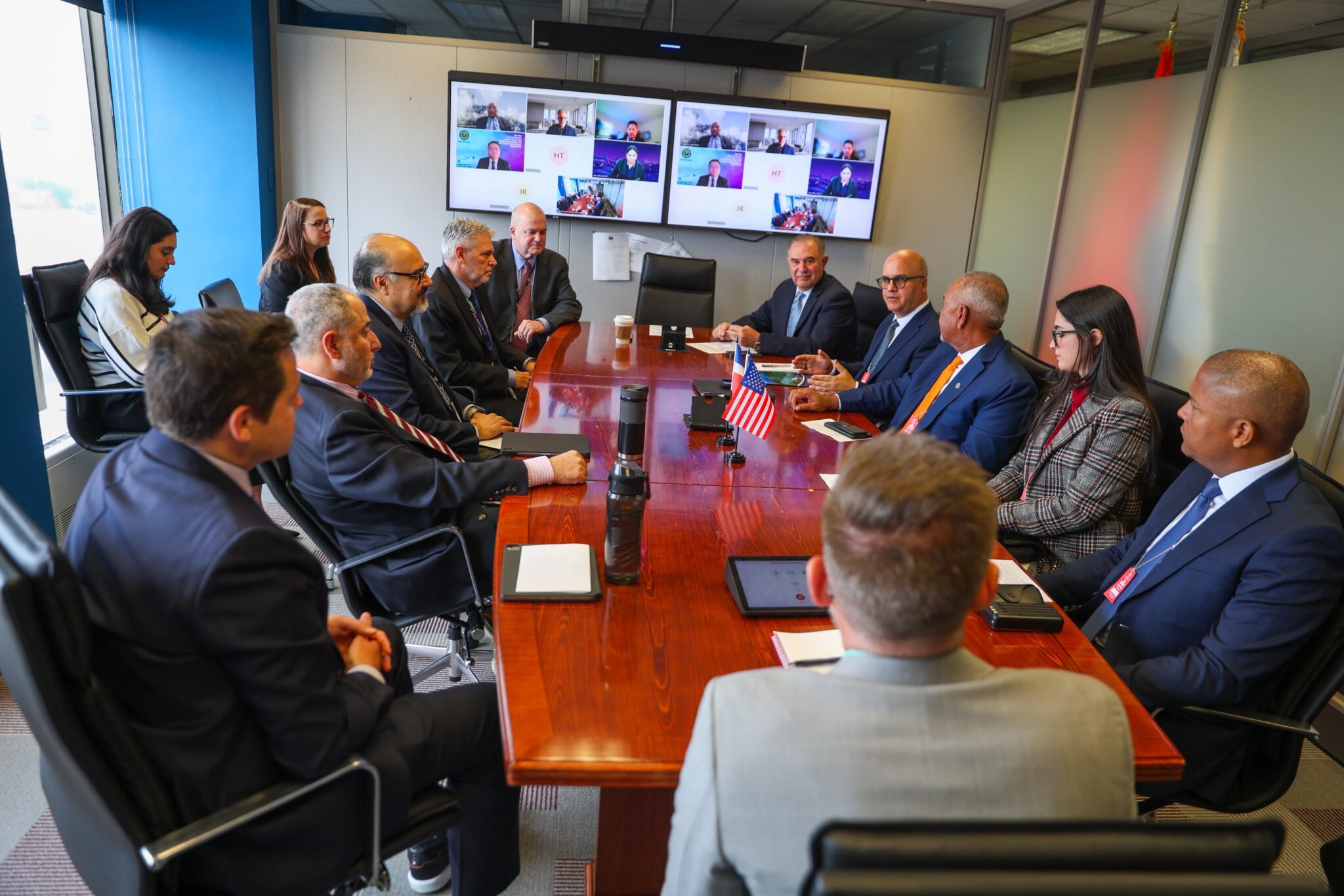 Participantes alrededor de una mesa con banderas y pantallas de videoconferencia.