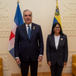 Dos personas posan entre las banderas de República Dominicana y Venezuela, en una sala con sofás.