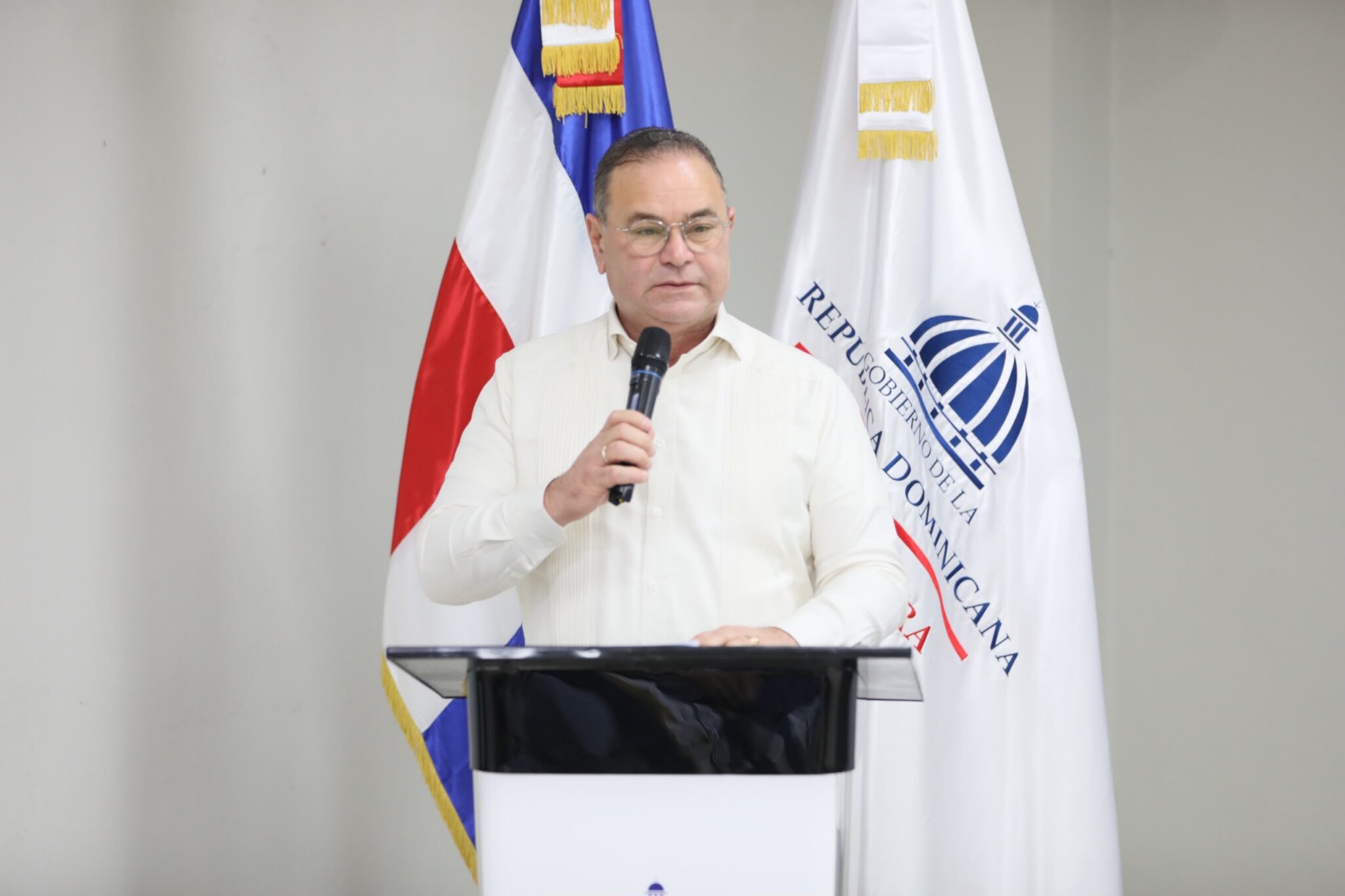 Persona con micrófono detrás de un atril, ante las banderas dominicana e institucional.