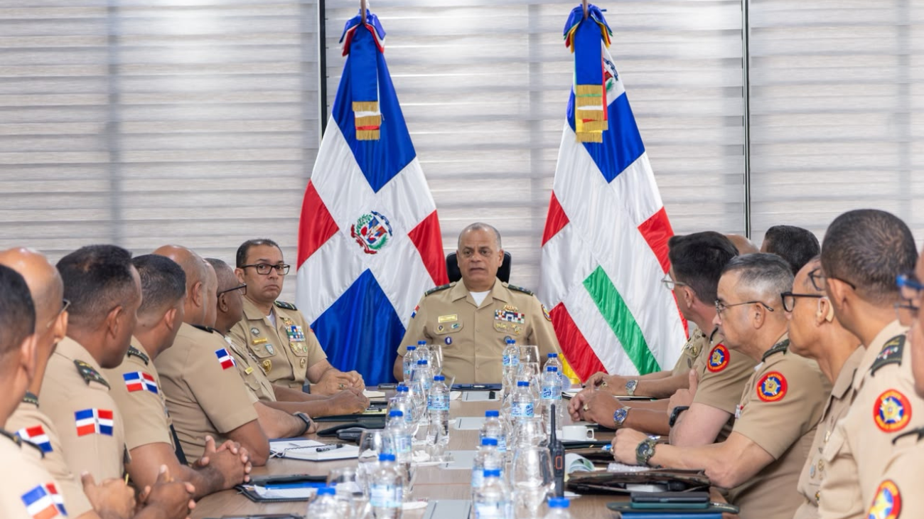 Militares uniformados se reúnen a ambos lados de una mesa, con dos banderas al fondo.