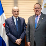 Dos personas con traje posan ante banderas en un despacho.