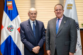Dos personas con traje posan ante banderas en un despacho.