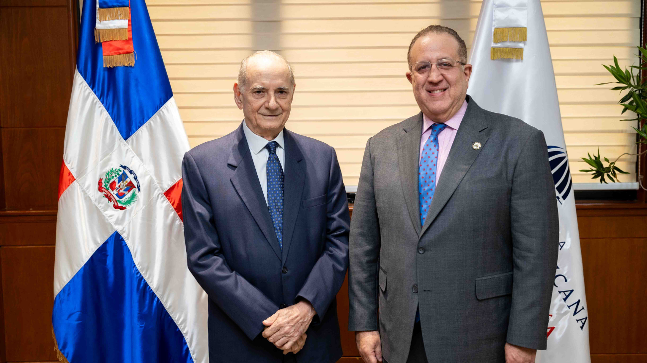 Dos personas con traje posan ante banderas en un despacho.