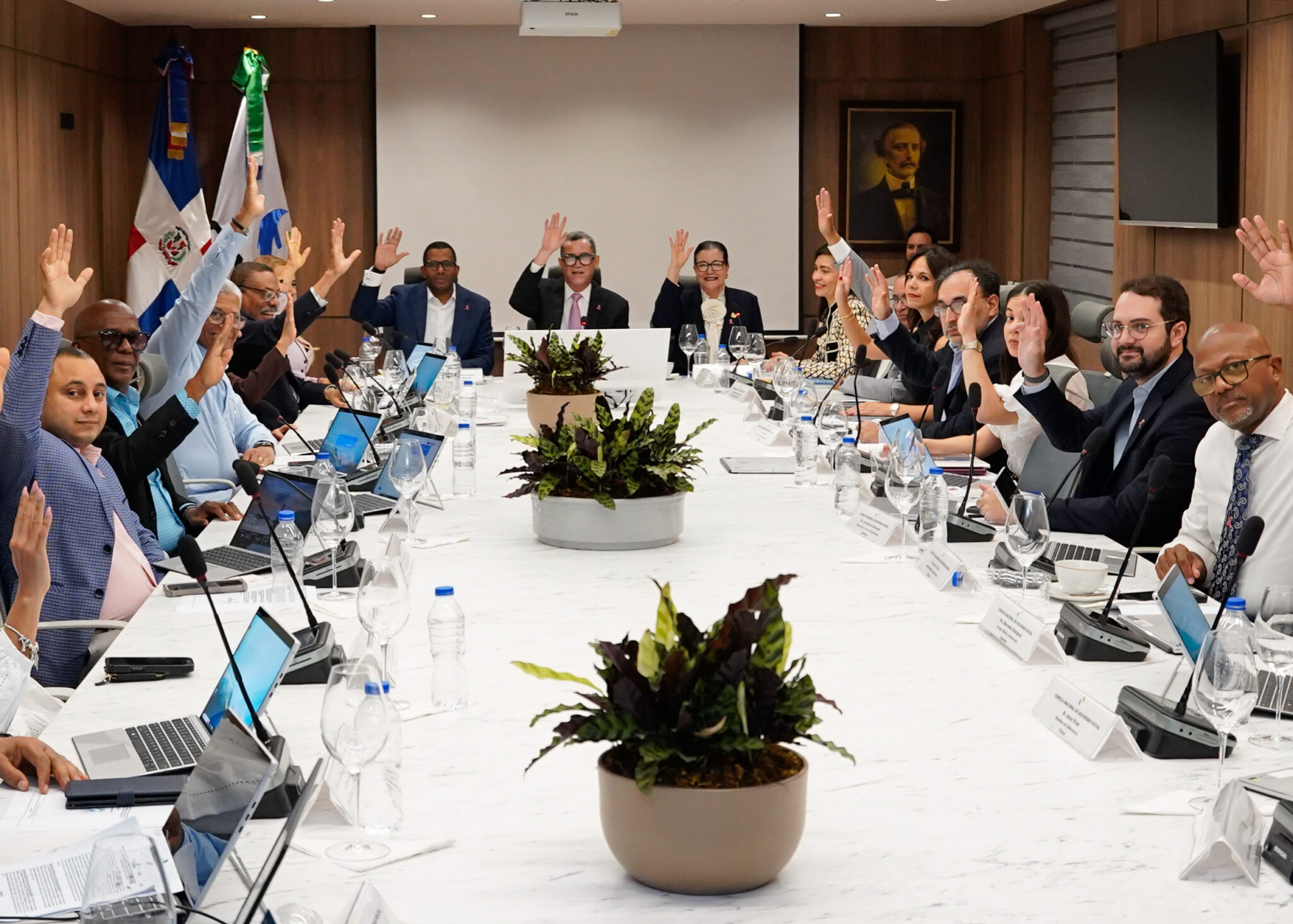 Participantes levantan las manos durante una reunión alrededor de una mesa.