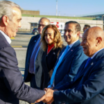 Varias personas con indumentaria formal se saludan en un exterior aeroportuario, con vehículos al fondo.