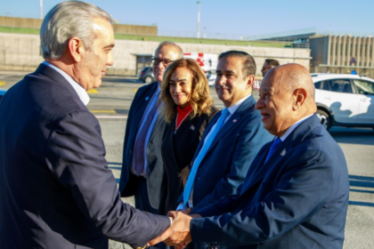 Varias personas con indumentaria formal se saludan en un exterior aeroportuario, con vehículos al fondo.