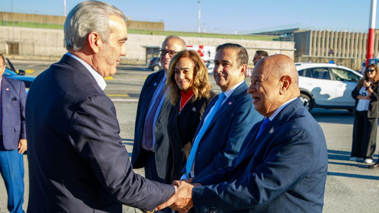 Varias personas con indumentaria formal se saludan en un exterior aeroportuario, con vehículos al fondo.