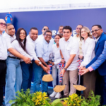 Participantes con palas y tierra ante un fondo institucional y plantas decorativas.