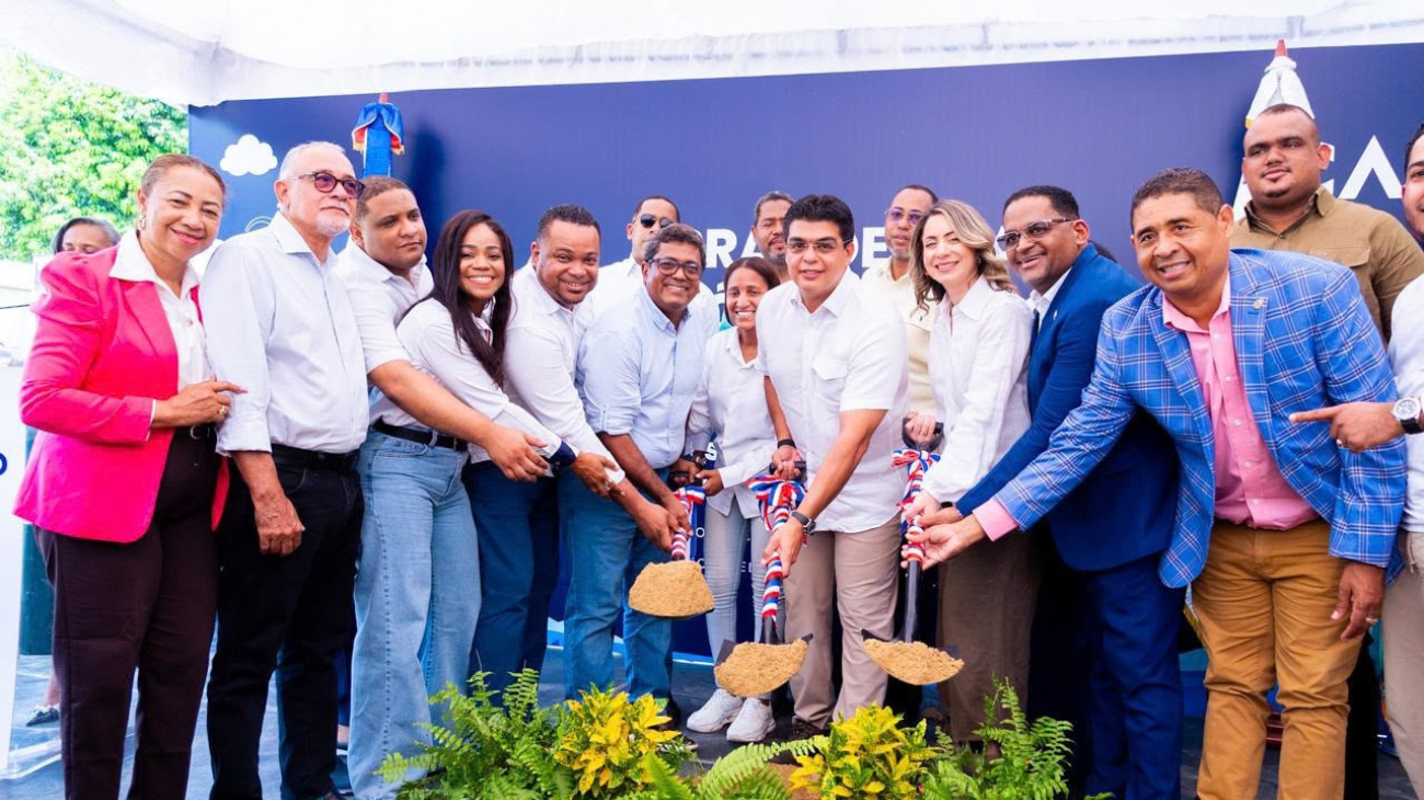 Participantes con palas y tierra ante un fondo institucional y plantas decorativas.