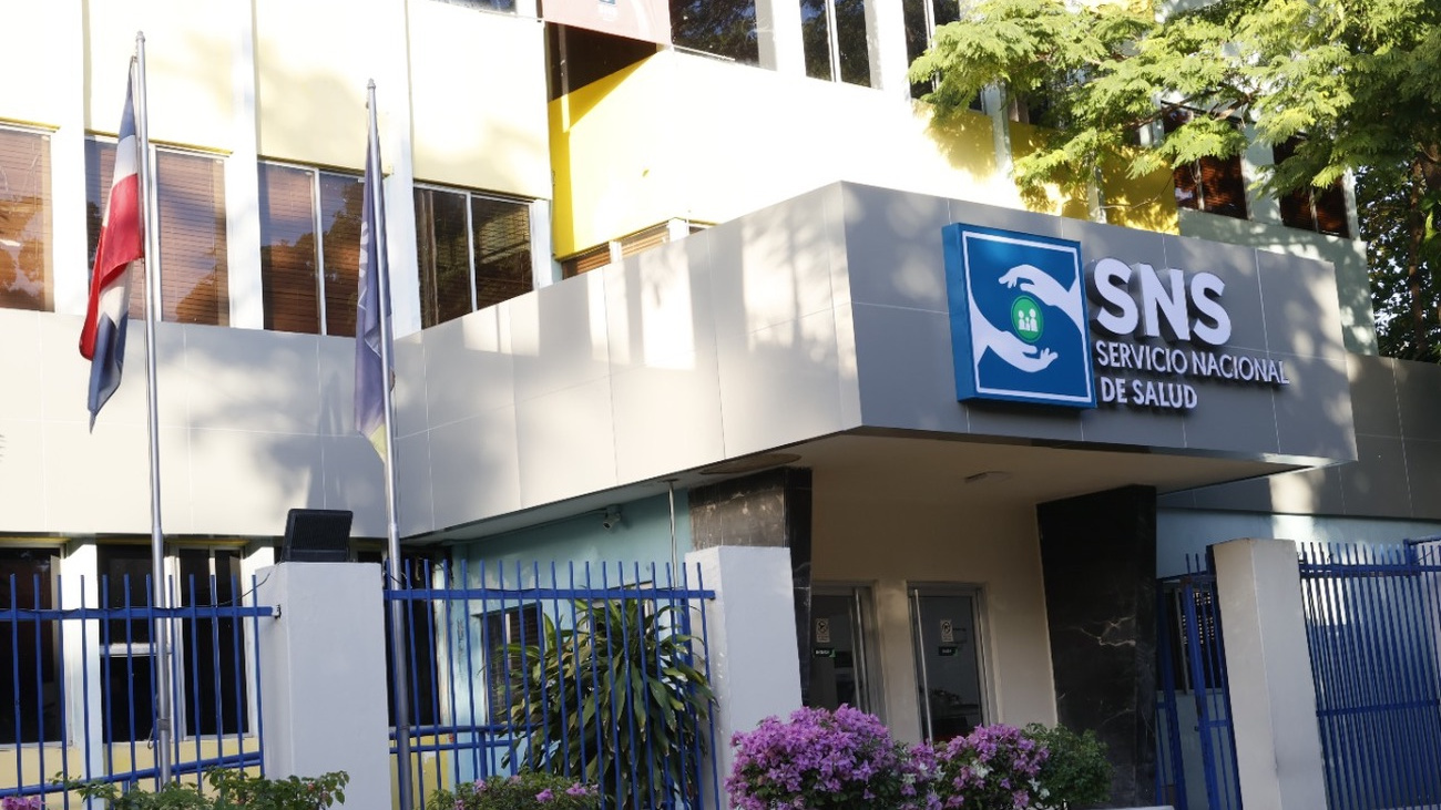 Fachada con letrero del Servicio Nacional de Salud, verja y banderas.