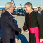 Saludo entre una persona con traje y otra con vestimenta clerical en un exterior aeroportuario.
