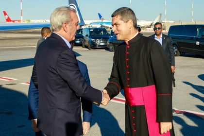 Saludo entre una persona con traje y otra con vestimenta clerical en un exterior aeroportuario.