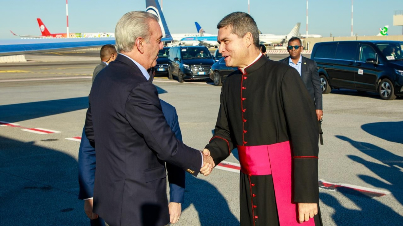 Saludo entre una persona con traje y otra con vestimenta clerical en un exterior aeroportuario.