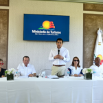 Participantes en una mesa y un ponente de pie, con pantalla del Ministerio de Turismo al fondo.