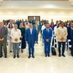 Personas de pie en filas de un auditorio, con indumentaria formal y algunas credenciales.