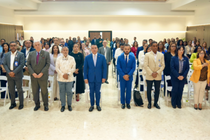 Personas de pie en filas de un auditorio, con indumentaria formal y algunas credenciales.