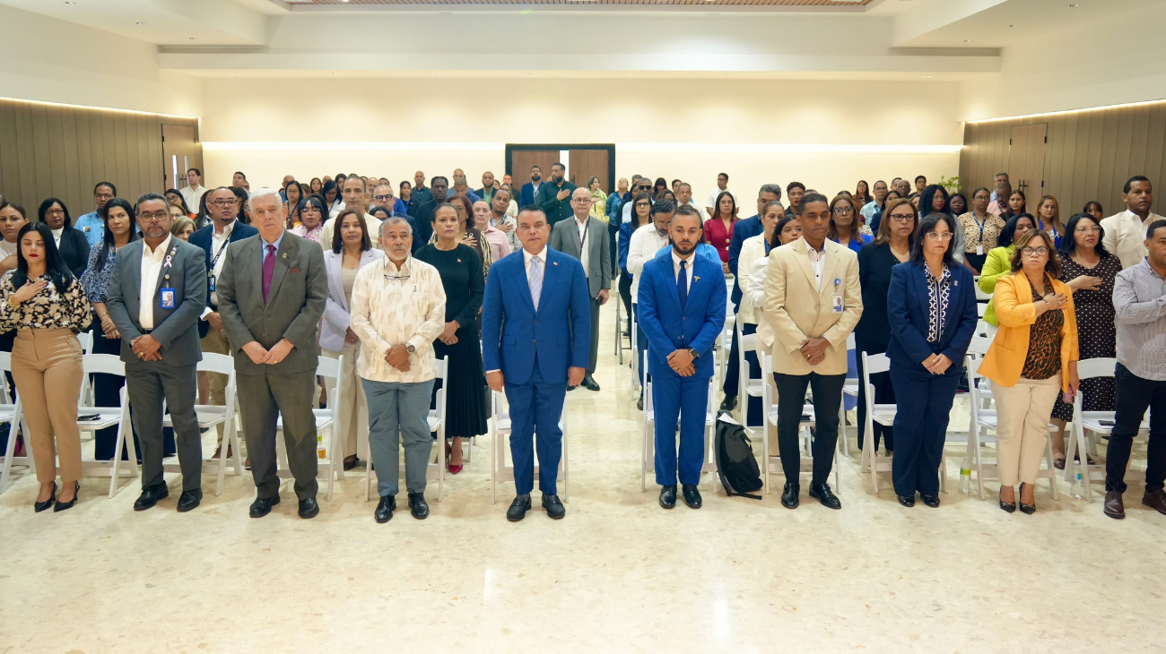 Personas de pie en filas de un auditorio, con indumentaria formal y algunas credenciales.