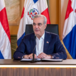 Persona ante un micrófono desde una mesa, con carpeta y banderas dominicanas detrás.