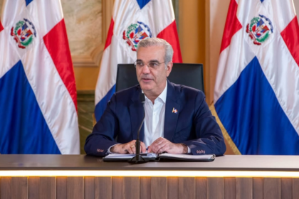 Persona ante un micrófono desde una mesa, con carpeta y banderas dominicanas detrás.