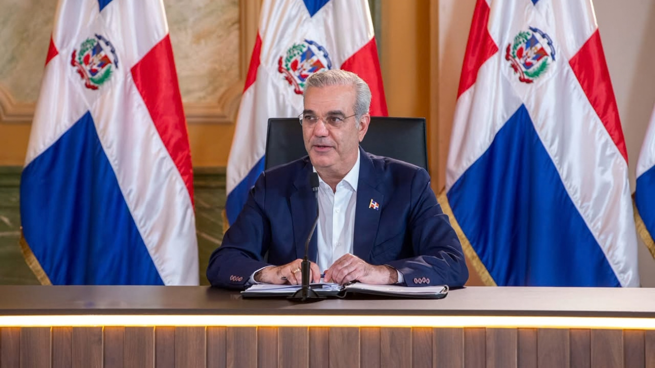 Persona ante un micrófono desde una mesa, con carpeta y banderas dominicanas detrás.