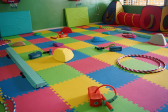 Aula vacía con suelo de espuma de colores, aros, túnel de juego y material de equilibrio.
